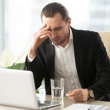 Stressed businessman having severe headache, holding painkiller remedy pill. Sick CEO with head cold, thinking about taking medicine with glass of water. Being ill at work, stress induced migraine.