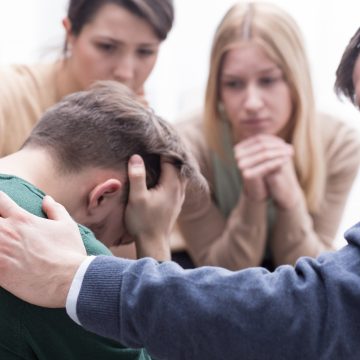 Close-up of a devastated young man holding his head in his hands and a group of friends in a supportive pose around him