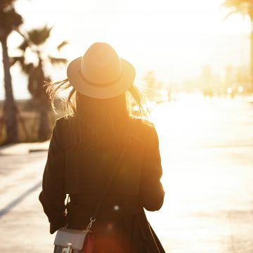 Back view of slender young woman wearing hat and coat walking down street on sunny spring day heading to city beach to enjoy beautiful sunset. People lifestyle travel and vacations concept