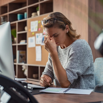 Exhausted businesswoman having a headache at office. Mature creative woman working at office desk feeling tired. Stressed casual business woman feeling eye pain while overworking on desktop computer.