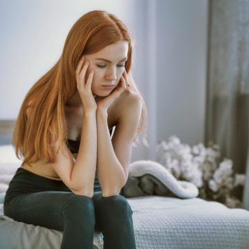 A teenager sits on the edge of her bed with her head resting in her hands. She is visibly upset and struggling with her body image.