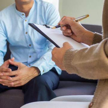 A patient sits across from a therapist during a treatment session for depression. The therapist is holding a clipboard and taking notes, while the patient sits across from the therapist, explaining their symptoms.